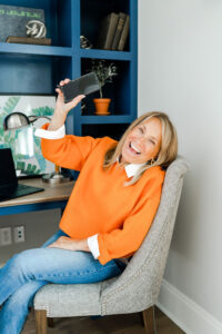 woman smiling at a desk while holding up an iphone