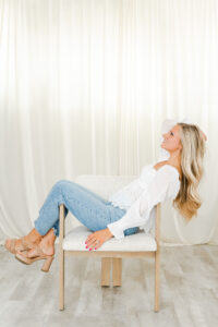 blonde woman in white shirt and jeans sitting sideways on chair with legs hanging off
