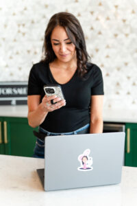 woman in black shirt looking at her phone while her laptop is in front of her