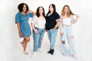 women in black, white, and denim smiling while posing together in front of a white wall during a bridal beauty branding session