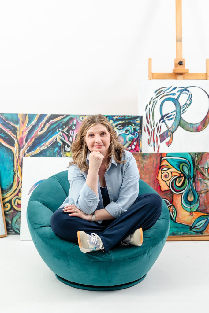 woman sitting in a teal chair with her artwork displayed behind her