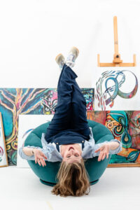 online coach lying upside down in a teal chair with her artwork displayed behind her