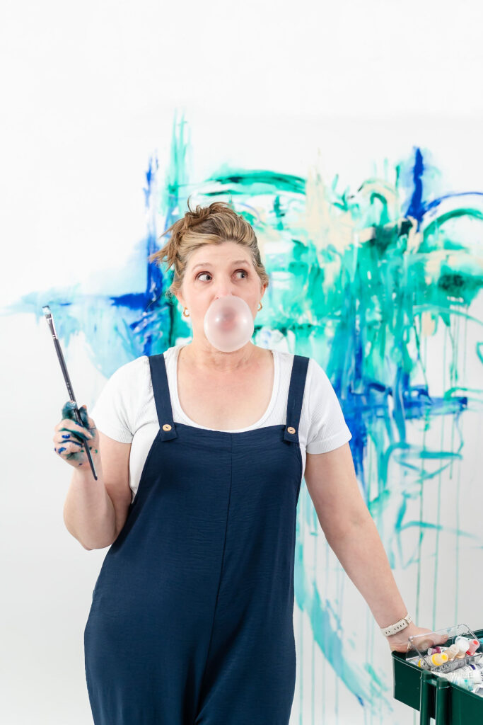 artist holding a paintbrush and blowing a bubble with her gum with her artwork in the background