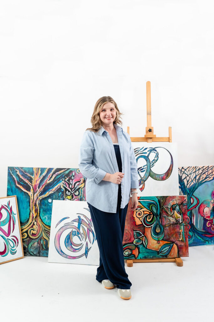 woman holding a canvas with artwork on it and multiple other pieces of art in the background