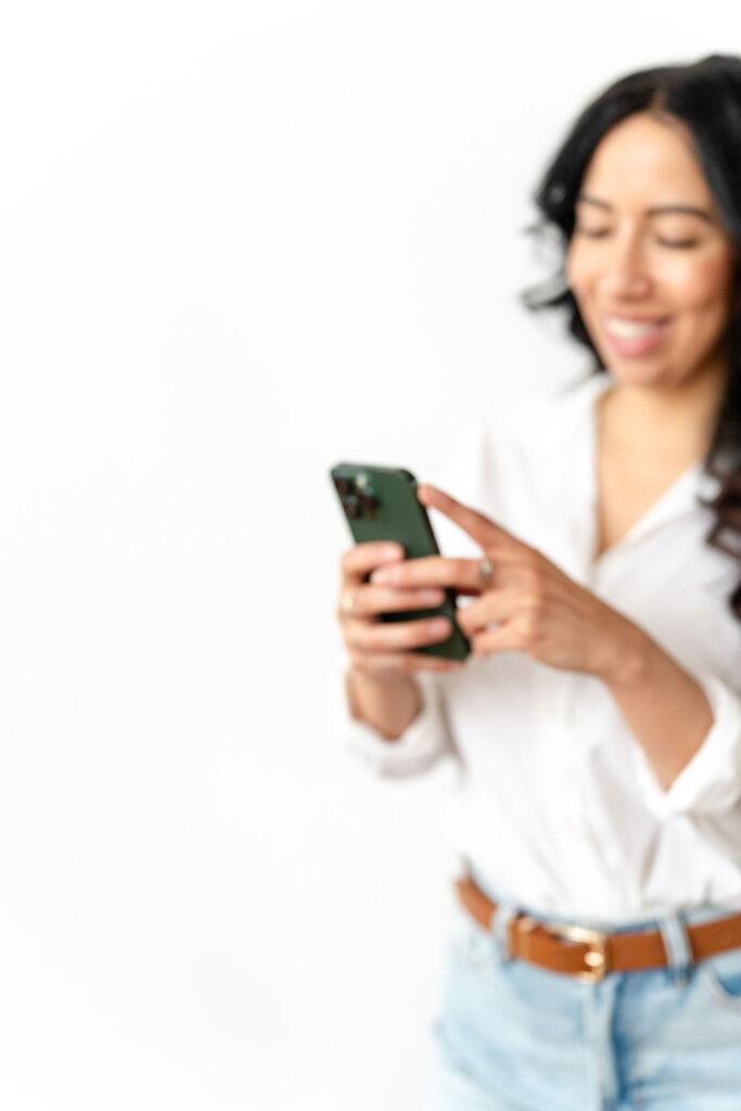 woman in white blouse and jeans standing up and smiling while holding her phone