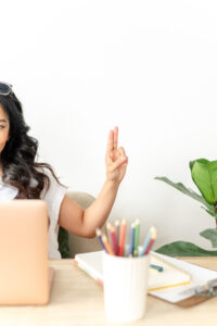 woman sitting at a table working on her computer and holding two fingers up in the air