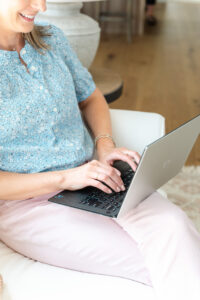 close up image of woman in blue shirt and pink pants sitting on a couch and typing on a laptop
