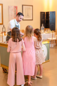 three girls standing next to the charcuterie cart watching a man make an order